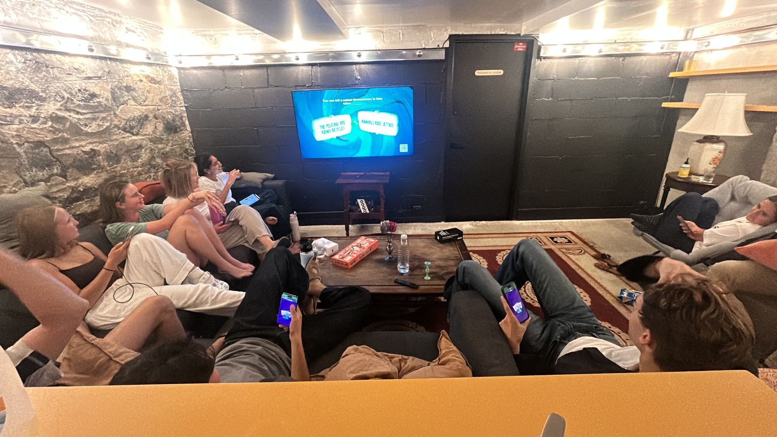 The basement lounge mid-game — everyone sprawled across the couches with their phones out, the Jackbox prompt up on the television against the black brick wall