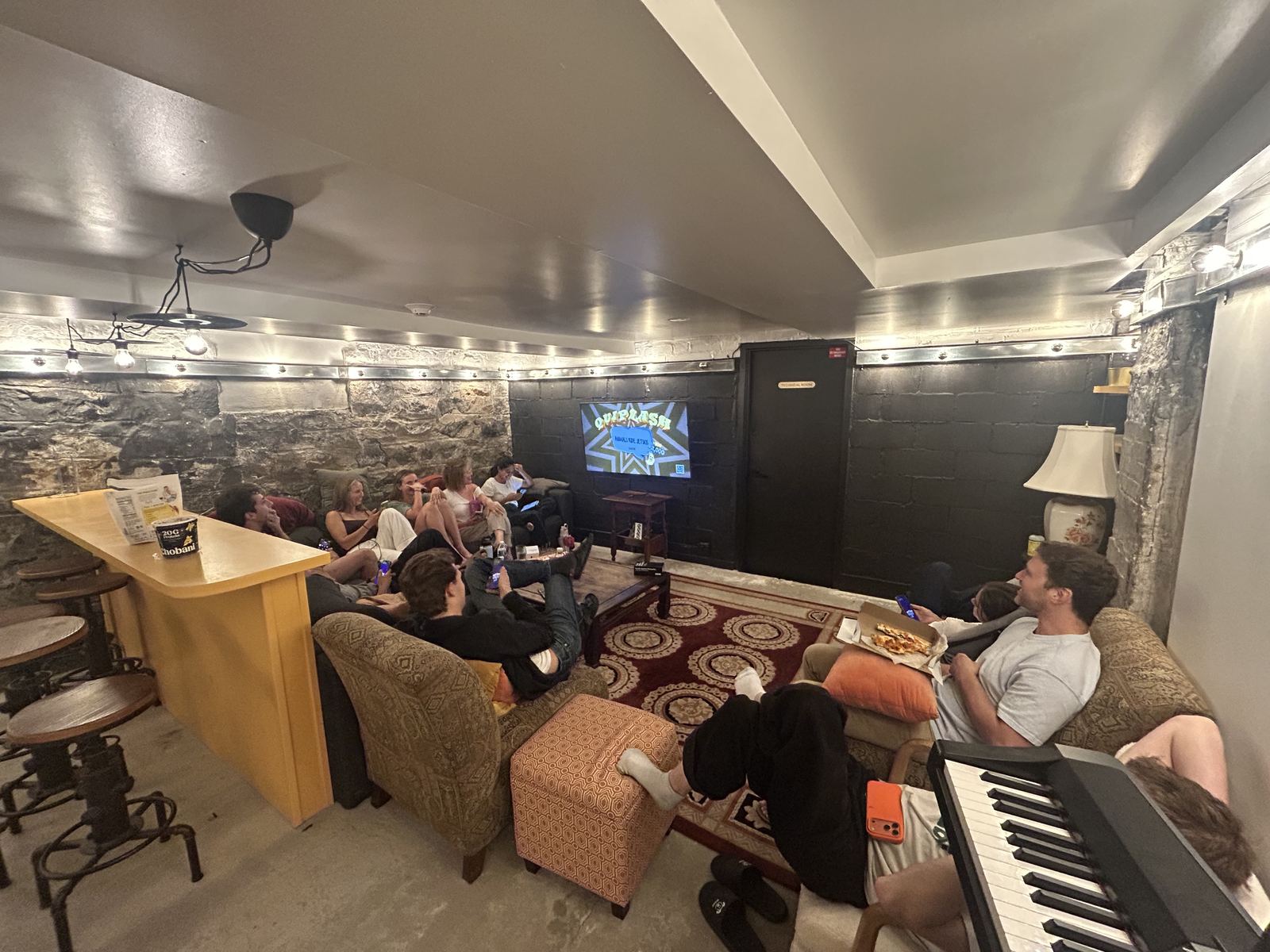 The wider room — stone wall and festoon lights, the burgundy rug, a keyboard in the foreground and the game still going on the screen