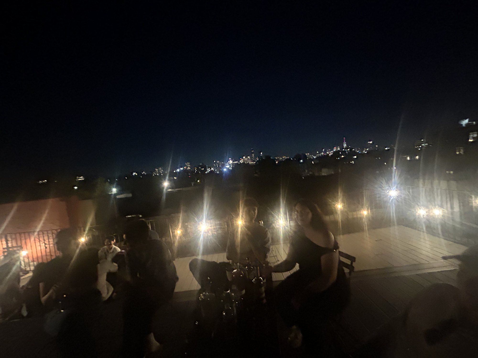 The wider roof after dark — people spread along the rail under string lights, the city glowing on the horizon