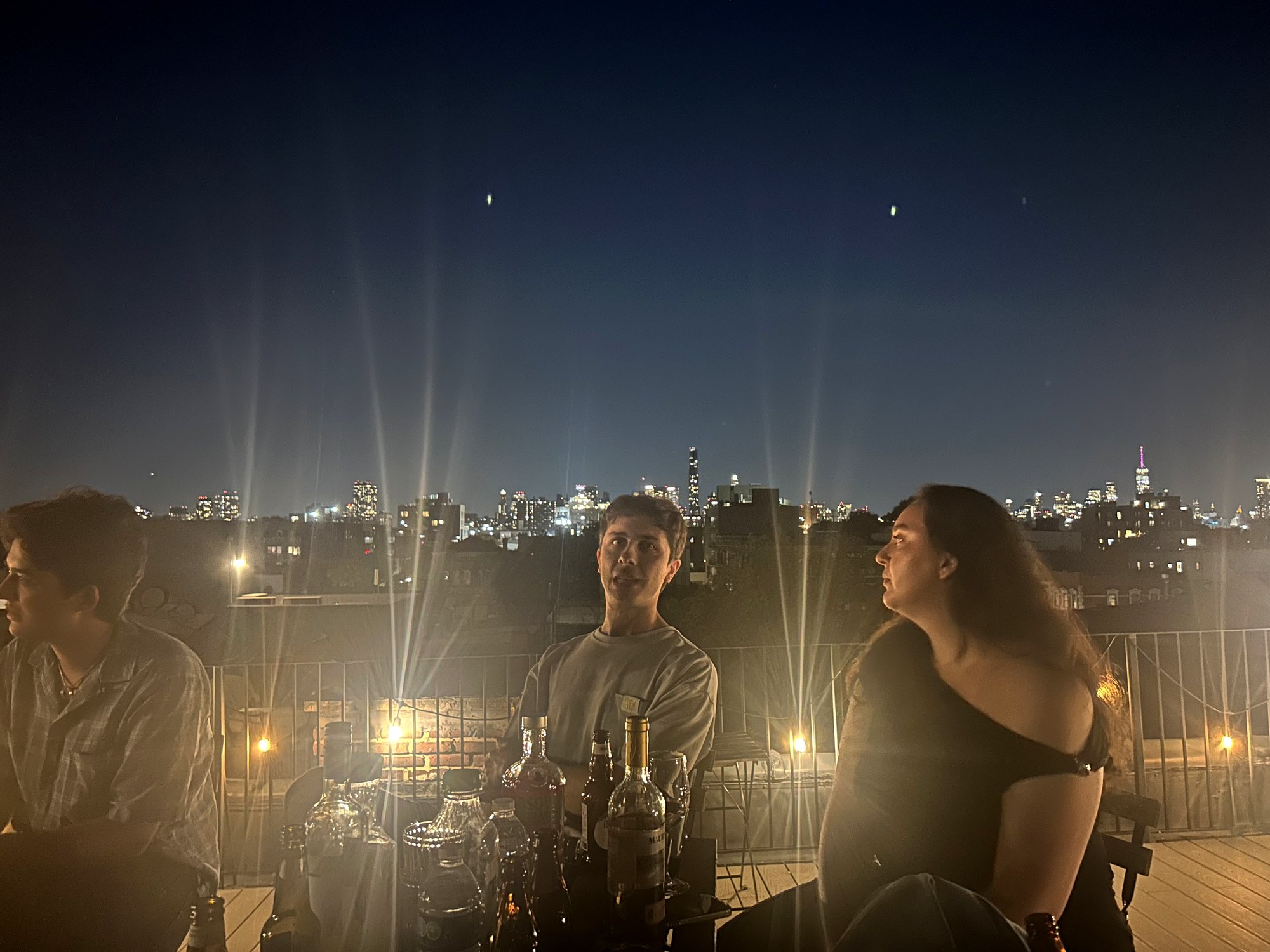 Three people around the table of bottles, the Manhattan skyline lit up behind them in the last of the blue hour
