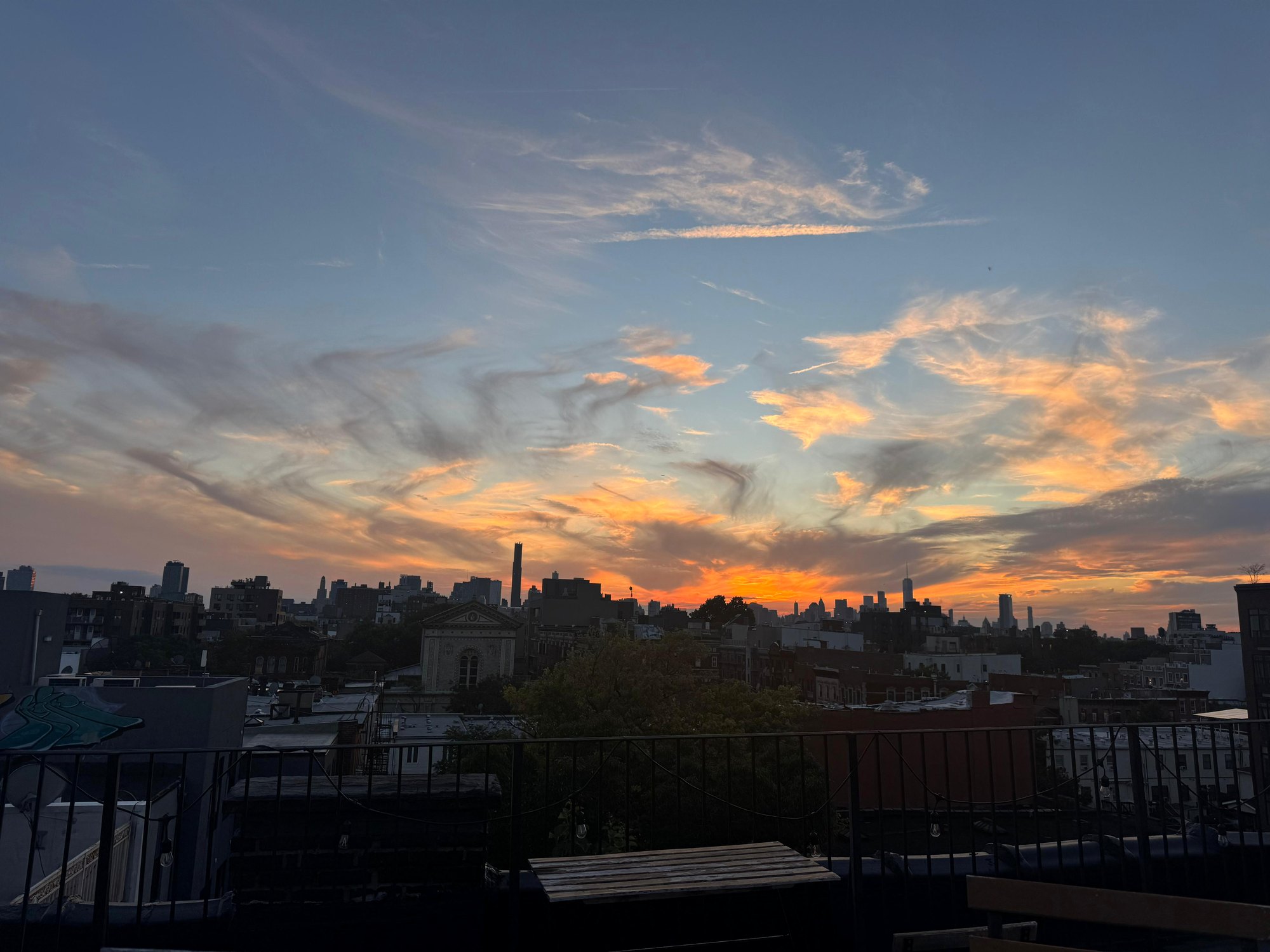 Sunset over the rooftops from the rail — orange light along the horizon behind the Manhattan skyline, string lights and the table in silhouette